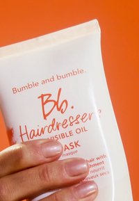 Vit tub med Bumble and bumble Hairdressers' Invisible Oil Mask med orange text och detaljer. Slät konsistens, hålls mot en orange bakgrund.