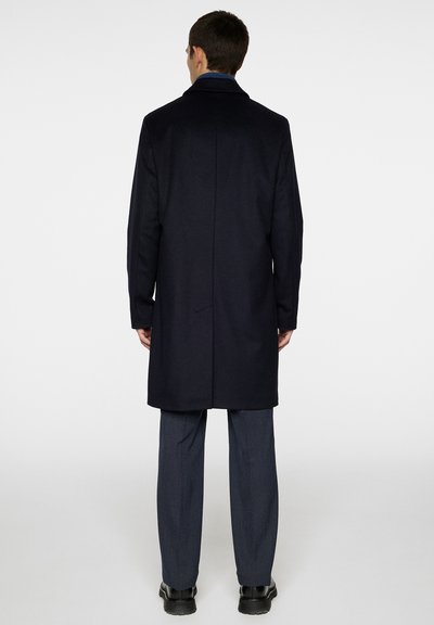 Mann, der mit dem Rücken zur Kamera steht, trägt einen dunklen Mantel, graue Hosen und schwarze Schuhe, vor einem schlichten hellen Hintergrund.