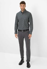 Ein Mann steht in einem dunkelblauen gemusterten Button-Up-Hemd, grauen Hosen mit einem schwarzen Gürtel und schwarzen Lederschuhen vor einem schlichten weißen Hintergrund.