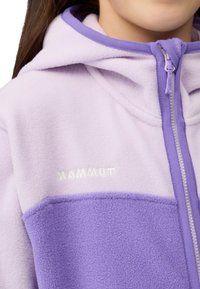 Giacca in pile di un leggero color lavanda con dettagli in un viola più scuro. Caratterizzata da un cappuccio, zip integrale e logo riflettente "MAMMUT" sul petto.