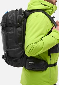 Sac à dos noir avec une finition texturée, des bretelles ajustables et plusieurs compartiments. Porté sur une veste verte éclatante avec une fermeture éclair à l'avant.
