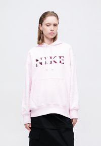 Rozā Nike džemperis ar kapuci, kangaroo kabatu un izteiksmīgu tumši sarkanu logotipu priekšā. Garas piedurknes ar ribotu aproci, brīva piegriezuma. Melns apakšdaļa.