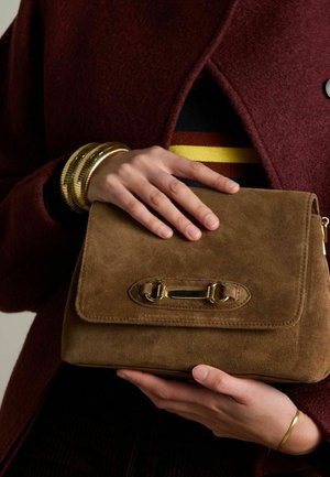 Borsa a tracolla in camoscio marrone con chiusura a patta, dettagli in oro e superficie testurizzata. Tenuta in mano, davanti a un indumento esterno color amaranto.