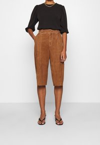Brown suede shorts with front pockets and stitching details, paired with a black top featuring puffed sleeves and a chain neckline.