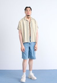Camicia a maniche corte beige a righe con bottoni e taschino, abbinata a pantaloni corti in denim azzurro chiaro e scarpe da ginnastica bianche.