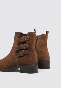 Botines de ante marrón que presentan una puntera cuadrada, tres hebillas decorativas y un cierre lateral con cremallera. El tacón es ancho y de color negro.