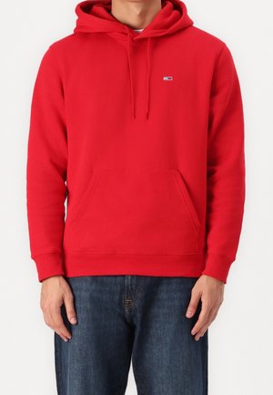 Mann trägt leuchtend roten Pullover-Hoodie mit Fronttasche und kleinem Logo auf der Brust, kombiniert mit dunkelblauer Jeans, stehend vor weißem Hintergrund.