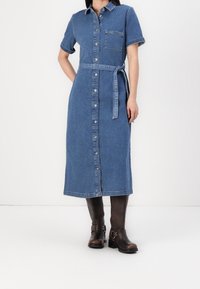 Sukienka koszulowa z denimu, krótki rękaw, kołnierzyk, zapięcie na guziki z przodu, taliowana z paskiem. Noszona z ciemnobrązowymi botkami sięgającymi kostek.