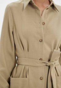 Robe chemise beige à col en tissu texturé avec une taille ceinturée, dotée de boutons et de poches avant.