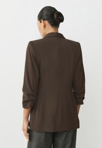 Sacou maro, croi pe măsură, cu mâneci lungi și ușor bufante, și guler tip rever. Material neted, design ajustat și buzunare cu fante.