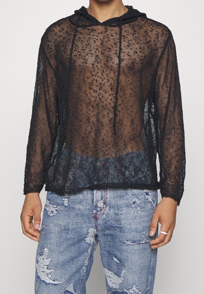 Personne portant un sweat à capuche noir transparent à manches longues et un jean bleu clair usé, debout avec les mains détendues le long du corps.