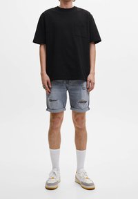 Camiseta de algodón negra con un bolsillo en el pecho, combinada con pantalones cortos de mezclilla azul claro con detalles desgastados y puños doblados. Zapatillas blancas.