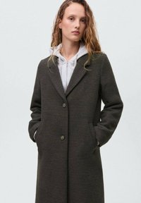 Young woman with curly hair wearing a dark gray buttoned coat over a light gray zip-up hoodie, hands in coat pockets, neutral background.