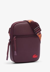 Borsa a spalla texturizzata viola con scomparto con zip, dotata di una tracolla con logo arancione e un logo accentuato rosso sul frontale.