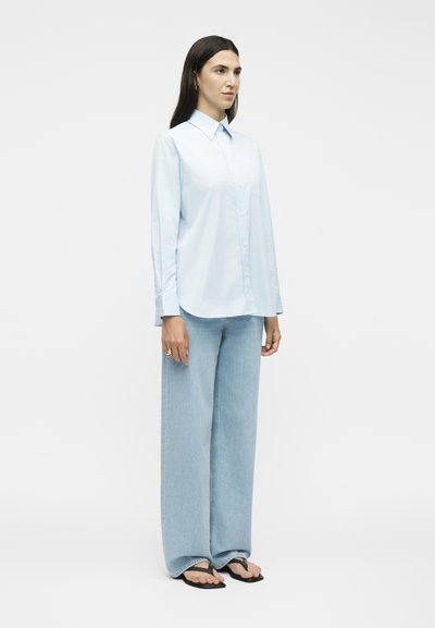 Γυναίκα με μακριά σκουρόχρωμα μαλλιά, που φοράει ένα ανοιχτό μπλε πουκάμισο με κουμπιά, φαρδιά ανοιχτό μπλε τζιν και μαύρα σανδάλια, στέκεται στραμμένη λίγο προς τα δεξιά.