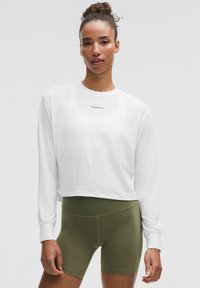 Valge pika varrukaga lühike topp "lululemon" logoga, mida on paari pandud oliivroheliste kõrge vöökohtadega lühikeste pükstega. Sile tekstuur ja mugav lõige.