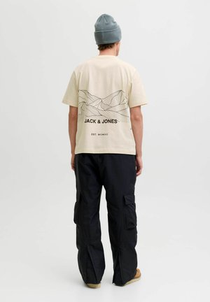 Beige T-Shirt mit kurzen Ärmeln und schwarzem Berg-Graphic sowie Markentext auf der Rückseite, kombiniert mit schwarzen Cargo-Hosen und einer grauen Mütze.