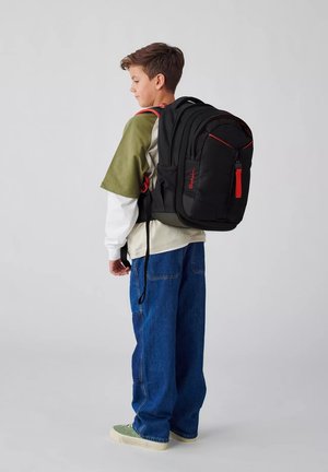 Schwarzer Rucksack mit roten Akzenten; gepolsterte Träger, mehrere Taschen, strukturiertes Material und ein Logo. Wird von einer Person in lässiger Kleidung getragen.