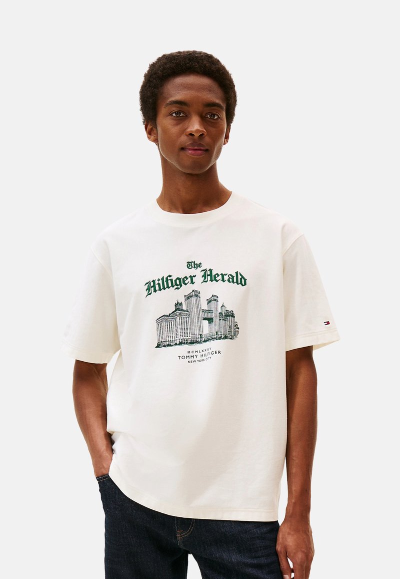 Joven vestido con una camiseta blanca de Tommy Hilfiger con un paisaje urbano verde y el texto "The Hilfiger Herald" impreso en el frente, combinada con jeans oscuros.