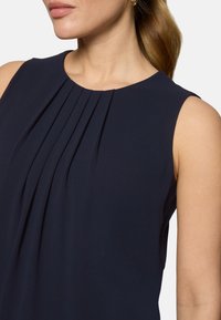 Femme portant un haut sans manches bleu marine avec des plis verticaux à l'avant, des boucles d'oreilles en anneau doré et les cheveux attachés en queue de cheval.