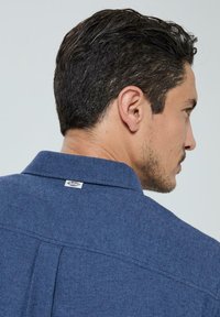 Chemise bleue à motif avec un tissu texturé, col boutonné et une petite étiquette au dos du cou. Coupée droite.