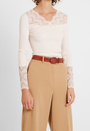 Mujer usando un top de manga larga color crema con encaje, metido dentro de pantalones beige de talle alto con un cinturón marrón texturizado.