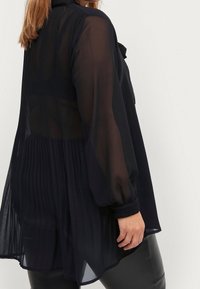 Femme portant un chemisier noir transparent à manches longues avec un dos plissé et un pantalon en cuir, debout devant un fond clair uni.