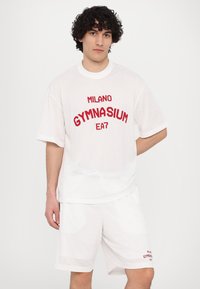 Baltos akių sportinės aprangos komplektas, kuriame yra trumpomis rankovėmis marškinėliai su raudonais užrašais "MILANO GYMNASIUM" ir derančios šortai su panašiu dizainu.