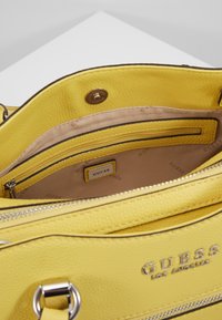 Keltainen Guess-olkalaukku, jossa on vetoketjupussukka ja beige sisävuori, johon on painettu "GUESS"-teksti sekä metallinen logo-levy.