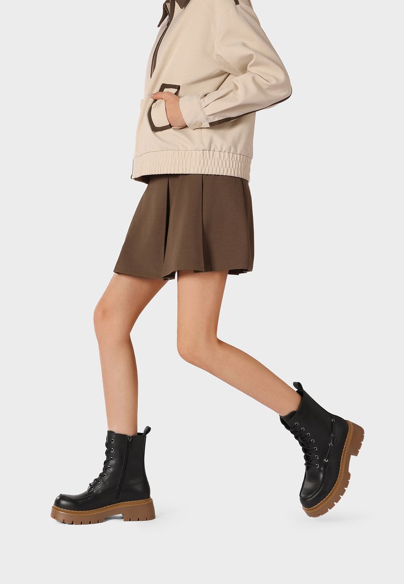 Persona caminando con botas negras con cordones y suelas marrones, falda marrón y chaqueta beige con la mano en el bolsillo.