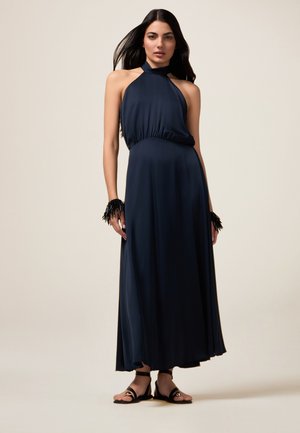 Marineblauwe halter maxi-jurk met een gathered taille, vloeiende stof en zwarte franje-accenten bij de polsen. Zwarte sandalen maken de look compleet.