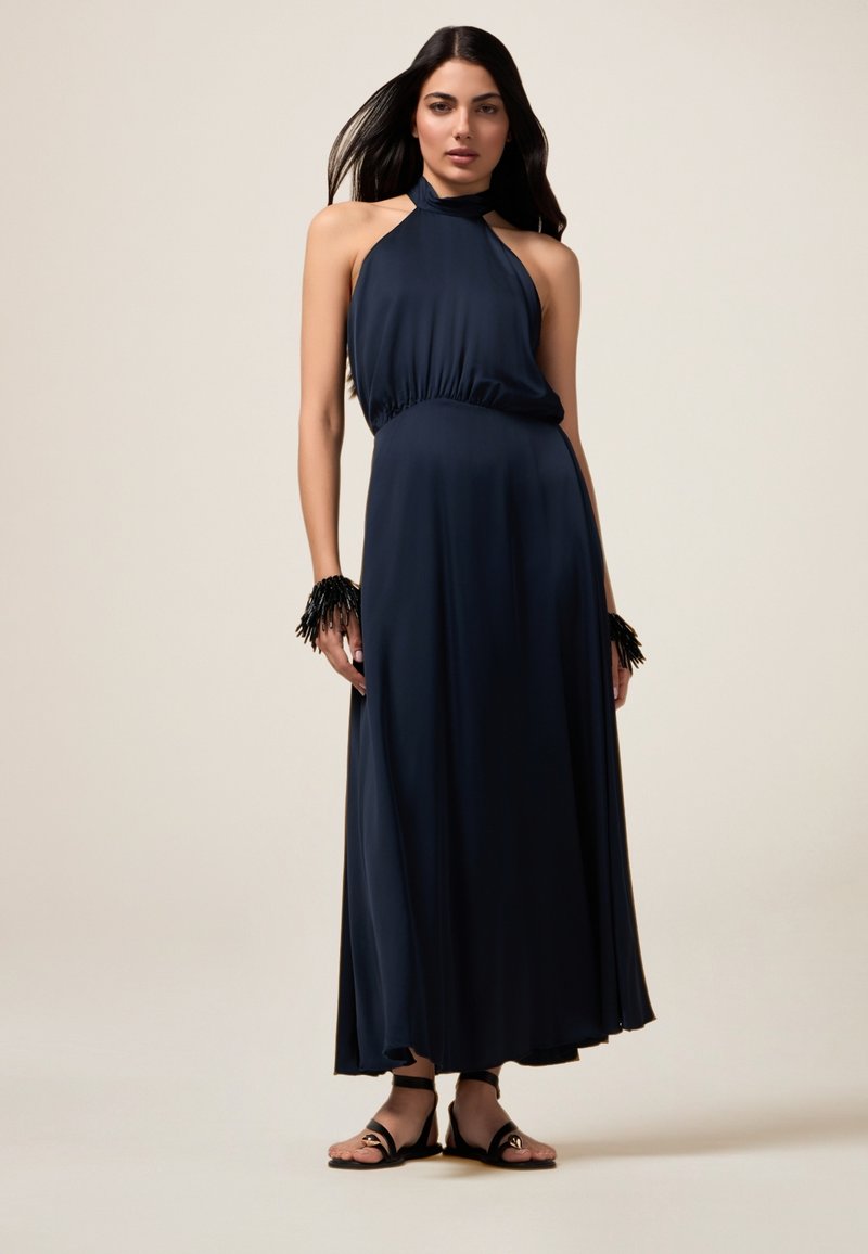 Navy Halter-Maxikleid mit gerafftem Taillenbereich, fließendem Stoff und schwarzen Fransenakzenten an den Handgelenken. Schwarze Sandalen runden den Look ab.