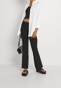 Bílá košile na knoflíky přes černý crop top, v kombinaci s černými zvonovými kalhotami a masivními černými platformovými sandály, držící malou kabelku.