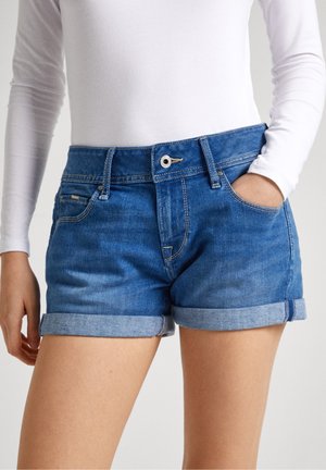 Personne portant un short en jean bleu retroussé et une chemise blanche à manches longues, main dans la poche, debout devant un fond neutre.