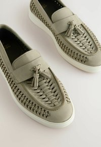 Lys grå læder slip-on sko med vævet detalje, flad hvid gummisål og dekorativ kvast på fronten.