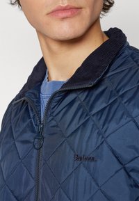 Marineblaue gesteppte Jacke mit einem Cordkragen, ausgestattet mit einem Reißverschluss und einem bestickten Etikett. Glatte Textur und Diamantmuster-Design.