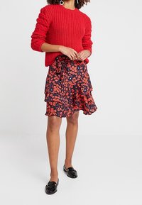 Punainen neulottu villapaita, jossa on rakennehihoja, yhdistettynä tummansiniseen kukalliseen volangihameeseen. Asusteena mustat tasapohjaiset kengät.