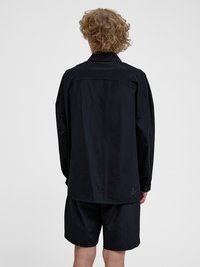 Persoon met krullend haar die een los zwart shirt met lange mouwen en een zwarte korte broek draagt, gezien van achteren tegen een effen lichte achtergrond.