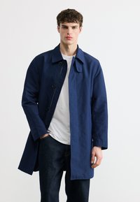 Νεαρός άνδρας με μπλε σκούρο παλτό πάνω από ένα λευκό μπλουζάκι και σκούρο τζιν, στέκεται με το ένα χέρι στην τσέπη του μπροστά σε ένα απλό φωτεινό φόντο.