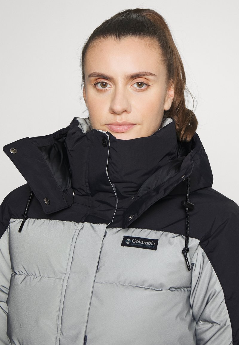 Puffjacke in Schwarz und Hellgrau, mit hohem Kragen, verstellbaren Kordeln und Columbia-Logo auf der Brust. Glatte Stofftextur.