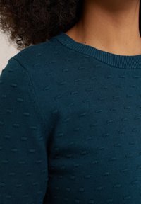 Pull tricoté couleur sarcelle avec un motif à pois texturé, un col côtelé et un tissu lisse. Gros plan sur le haut du corps.
