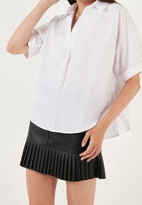 Weißes kurzärmliges Hemd mit Knöpfen und Kragen, kombiniert mit einem schwarzen gefalteten Leder-Minirock, der oben glatt und unten strukturiert ist.