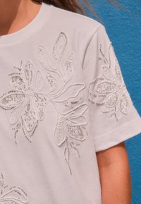 Biele bavlnené tričko s krátkym rukávom zdobené kvetinovou výšivkou so striebornými korálkami a flitrami. Textúrovaný dizajn na oboch rukávoch.