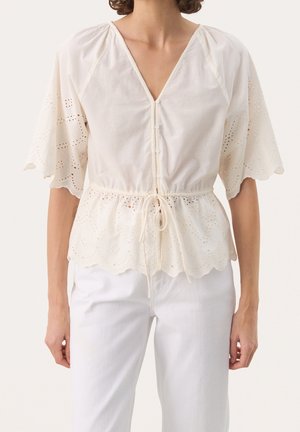Femme portant un chemisier en dentelle ajourée blanche à manches courtes, bords festonnés, boutons à l’avant et taille à cordon, associé à un pantalon blanc.