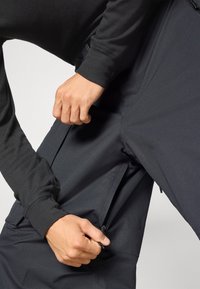 Čierne, vodeodolné nohavice s hladkým povrchom a bočnými vreckami na zips. Ruka sa snaží upraviť zips vrecka.