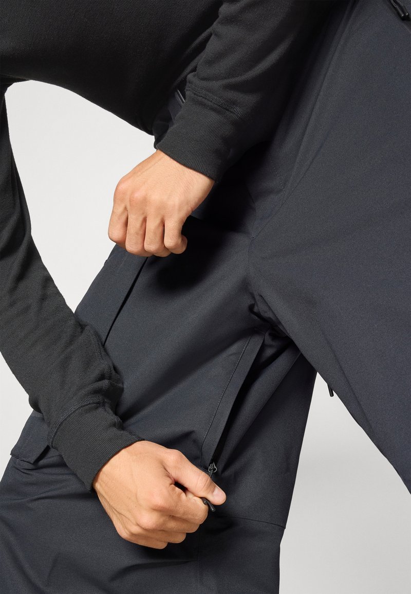 Black, water-resistant pants, featuring a smooth texture and zippered side pockets. A hand reaches to adjust the pocket zipper.