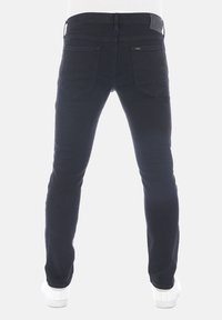 Vaqueros de denim negro vistos desde atrás, con un corte ajustado, cinco bolsillos y una etiqueta de cuero en la cintura. Combinados con zapatillas blancas.