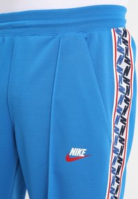 Modré sportovní kalhoty s měkkou texturou, s bílým logem Nike a vzorovaným bočním pruhem v černé, červené a modré barvě.