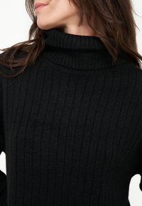 Woman wearing a black ribbed knit turtleneck sweater, showing neck and part of the face turned sideways.
