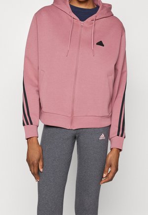 Osoba nosząca różową bluzę z kapturem na suwak z czarnymi paskami na rękawach oraz szare spodnie Adidas z logo na lewej nodze.